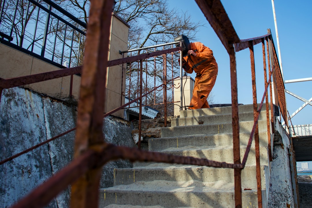 A worker in an orange protective suit and welding mask is seen welding a rusty metal railing on a concrete staircase under a clear blue sky. The composition, taken from a low angle through the railing, emphasizes the texture and weathering of the environment, giving the scene an industrial, gritty atmosphere.